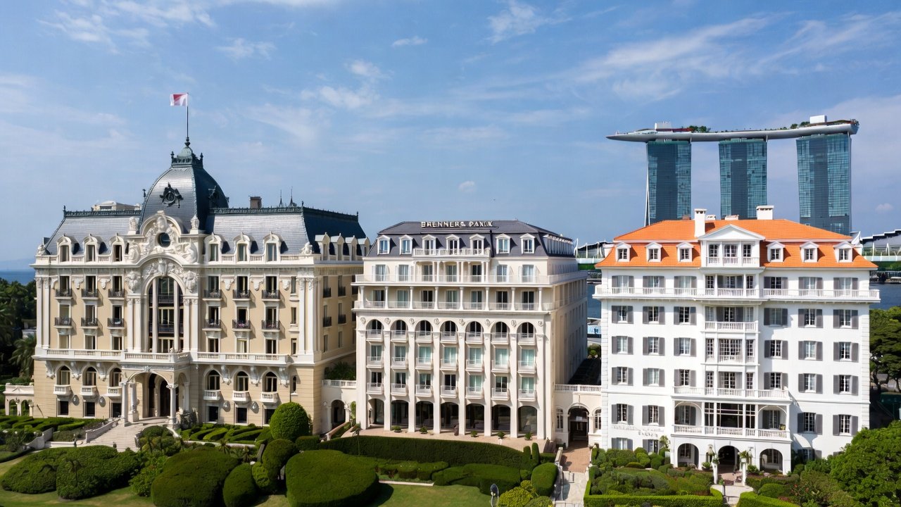 Three grand hotel buildings above clipped gardens under a blue sky, with modern towers in the distance